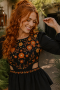 A smiling woman with long wavy red hair wears a black long-sleeve dress with orange and yellow floral embroidery across the bodice and a smocked waistband. The photo is taken outdoors in a garden-like setting.