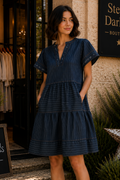 A woman wearing a dark blue tiered mini dress with short puff sleeves and pockets, standing in front of a boutique entrance with a sign that reads 'Stella, Darling Boutique'.
