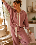 A woman modeling a dusty rose two-piece loungewear set consisting of a long-sleeve hooded sweatshirt and matching sweatpants with cream-colored drawstrings, posed in a bright, modern boho-style living room.
