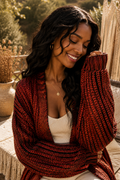 A woman wearing a chunky knit open-front cardigan in a deep rust red and black pattern, layered over a cream ribbed top. She is sitting outdoors on a woven bench with warm natural light and boho decor in the background.